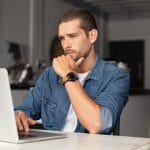 Serious young man working on laptop at home, reading about blog post length for his therapy website's SEO. Represents how private practice seo supports therapists in boosting their rankings on search engines and attracting their ideal clients.