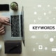 Photo of a person's hands using their computer next to a graphic of the word keywords in a search bar. This photo represents the way counselors can use mental health keywords and counseling keywords to improve their SEO through blogging.