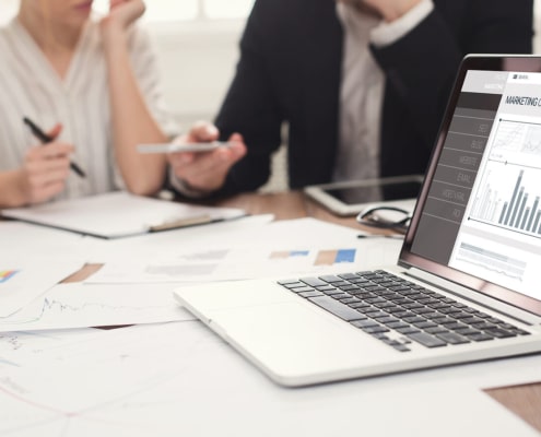 Photo of a laptop with data pulled up and two employees talking in the background. Represents how private practice seo benefits therapists who are too busy to learn themselves. They can have a team member learn and still benefit.