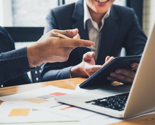 Two people work on laptop and tablet with papers on a table. Want to learn the best healthcare SEO strategy? Work with us today to make healthcare SEO work for you!