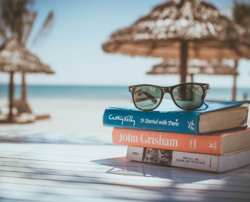 Shows a beach scene with a stack of books that have sunglasses on them. Represents how seo for mental health can help therapists with the summer slump.