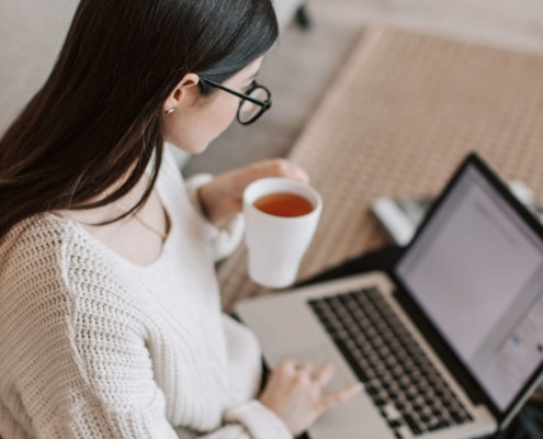 A woman in glasses and a cozy sweater works on her laptop while holding a cup of tea, symbolizing search engine optimization for counselors and SEO marketing for therapists.
