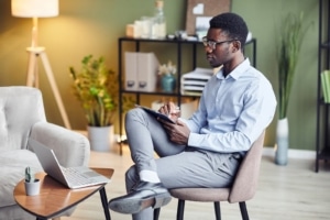 Image of a professional African American man sitting in a chair writing while looking at a laptop. Discover how to elevate your website’s performance and find your ideal clients with SEO consulting.