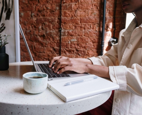 A person typing on a laptop at a café table, working on content strategy. This represents an SEO copywriting service and the importance of copywriting blogs for SEO to improve online visibility.